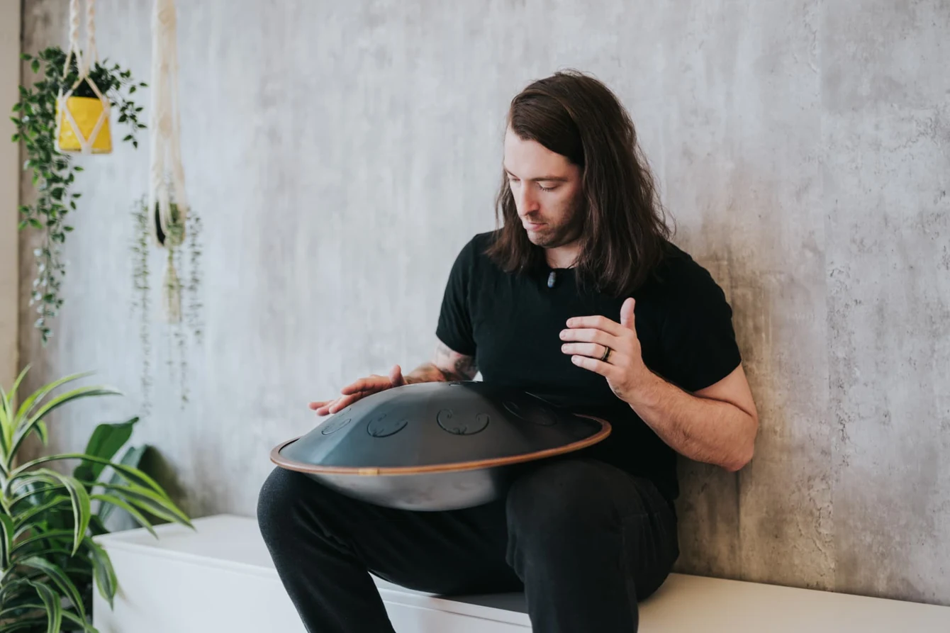 Travis seated beside plants, playing a handpan